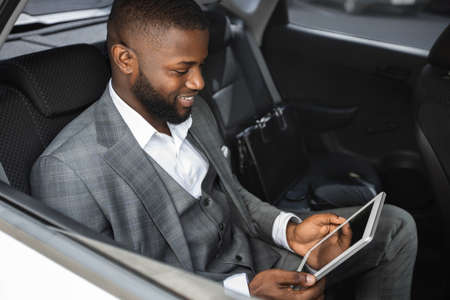 Smiling black guy businessman holding digital tablet with blank screen in luxury car. African american cheerful man using business application on pad while having business trip by car, copy spaceの写真素材