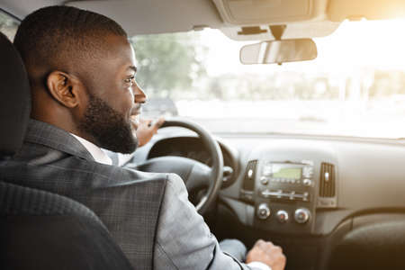 Closeup of smiling african american businessman in silver suit driving car, shot from behind. Happy bearded black man driver holding hands on wheel and gear, driving luxury car, looking at empty spaceの写真素材