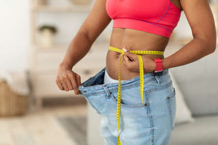 Slimming. African Woman After Weight Loss Measuring Thin Waist Wearing Oversize Jeans Standing Indoors, Cropped.の写真素材