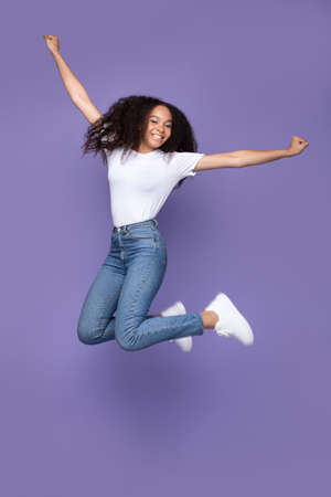 Happy African American Woman Jumping Posing In Mid-Air Over Purple Studio Background, Smiling To Camera. Happiness, Joy And Positive Emotions Concept. Full Length, Vertical Shotの写真素材
