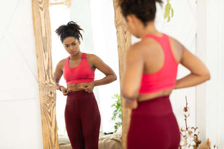 Unhappy African American Woman Touching Fat Belly Standing Near Mirror, Gaining Kilograms And Not Losing Excess Weight Dieting At Home. Unsuccessful Slimming Concept. Selective Focusの写真素材