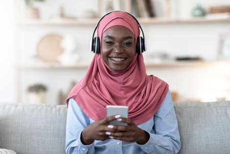 Black muslim girl using smartphone and wireless headphones at home, listening music, enjoying playlist or melodies app, wearing braces and pink veil, smiling at camera, copy spaceの写真素材