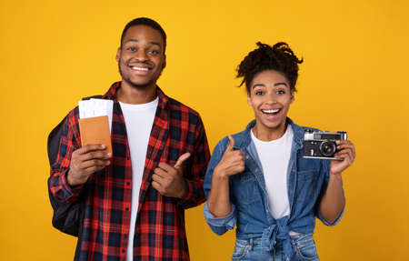 Happy Black Couple Holding Photo Camera And Travel Tickets Standing On Yellow Background. Studio...