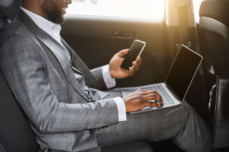 Unrecognizable black businessman using smartphone and laptop in car on a way to office. African american manager working on laptop, chatting with clients on smartphone, cropped, side view, sun flareの写真素材