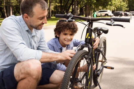 Closeup portrait of cheerful dad and son fixing bike using wire cutter at public parkの写真素材