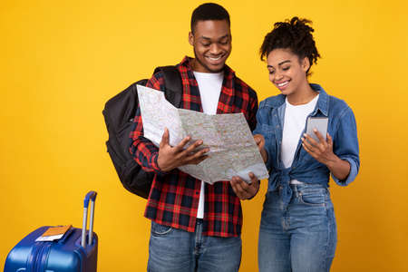 Happy Black Tourists Couple With Smartphone And Map Choosing Destination For Vacation Standing Over Yellow Background In Studioの写真素材