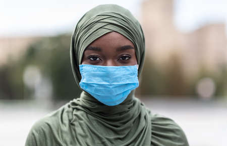 Portrait of black muslim woman in hijab wearing medical protective face mask outdoors on street for...