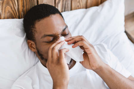 Sick African American Guy Blowing Runny Nose In Paper Tissue Lying In Bed At Home. Ill Black Man...