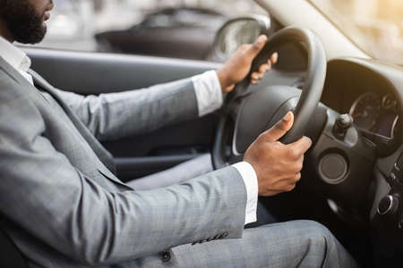 Closeup of unrecognizable black businessman in silver suit driving car, african american entrepreneur going to business meeting by his own car, holding both hands on steering wheelの写真素材
