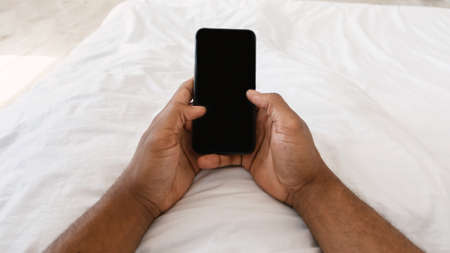 Unrecognizable African Man Using Smartphone With Blank Screen, Holding Cellphone Lying In Bed At Home. Mobile Application, Texting On Phone, And Online Chat Concept. Panorama, Cropped, POVの写真素材