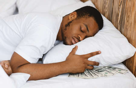 Black Man Sleeping Hiding Money Savings In Cash Under Pillow Resting Lying In Bed At Home. Wealth, Financial Safety And Literacy, Banking Services Concept. Selective Focusの写真素材