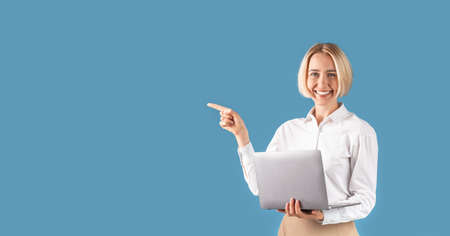 Happy business lady with laptop computer pointing aside at space for your text, blue studio background. Smiling female entrepreneur presenting or advertising something. Panoramaの写真素材