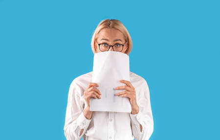 Shocked female entrepreneur peeking from behind documents over blue studio background, panorama....