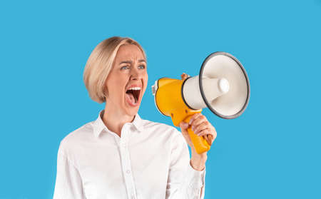 Agitated blonde woman in formal wear shouting into megaphone on blue studio background, panorama....