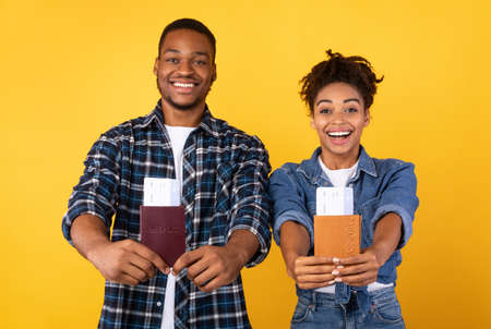 Vacation Concept. Joyful Black Tourists Couple Showing Travel Flight Tickets To Camera Posing Standing Over Yellow Studio Background. Joy Of Traveling, Holiday Trip And Tourismの写真素材