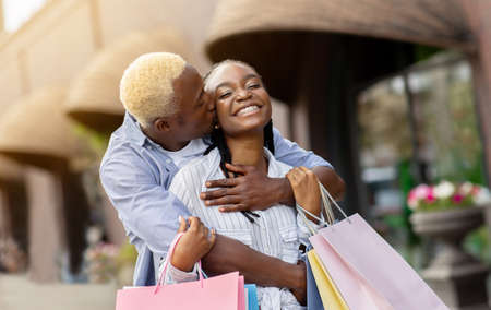 Hugs on walk at shopping. Cheerful african american guy kissing girlfriend with colored bags near shops, sun flare, free spaceの写真素材