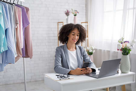 Clothes designer, seller and shopper consultant. African american woman typing on smartphone near laptop in studio interior at home, copy spaceの写真素材
