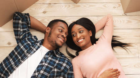 Black millennial woman with her boyfriend lying among carton boxes on floor of their new house, top...