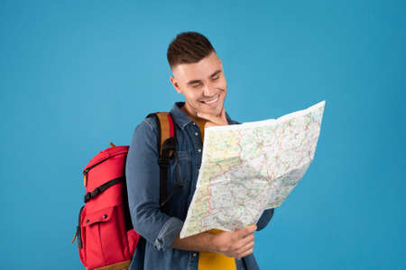 Portrait of happy young traveler with backpack and map on blue background. Positive millennial hiker...