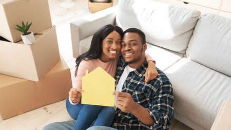 Black millennial guy with his girlfriend hugging and holding paper house while sitting on floor of...