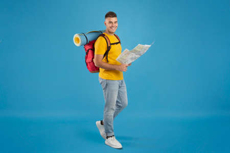 Full length portrait of positive young tourist with camping equipment and map walking on blue studio...