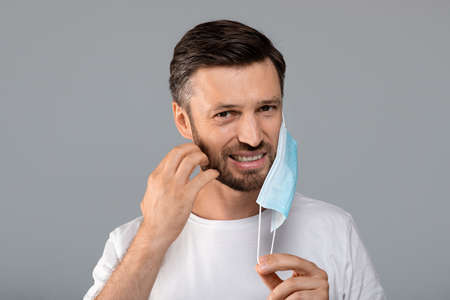 Bearded man moved protective medical mask from face to scratch beard, grey studio background, free...