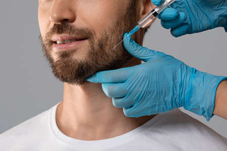 Unrecognizable bearded man getting beauty injection in salon, closeup. Cosmetologist hands in rubber gloves doing anti-aging procedure for man over grey studio background. Male cosmetology conceptの写真素材