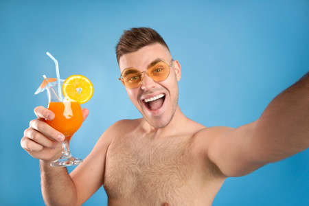 Excited young man in sunglasses holding refreshing summer cocktail and taking selfie on blue studio background. Funky guy enjoying his tropical summer vacation, making photo of himself on beach partyの写真素材