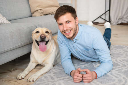 Happy Owner. Cheerful positive guy lying on the floor with his golden retriever and looking at camera. Happy man relaxing on carpet, posing with pet companion in modern apartment near sofaの写真素材