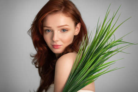 Natural Beauty. Red-Haired Young Lady Posing With Green Plant Leaves Standing Over Gray Studio...