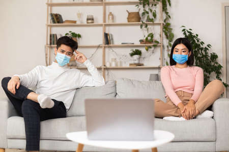 Coronavirus Self-Isolation Quarantine. Asian Couple In Medical Masks Watching Movie Sitting On Sofa...