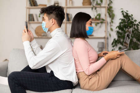 Japanese Couple In Protective Masks Using Cellphone And Laptop Sitting Back-To-Back At Home. Distant...