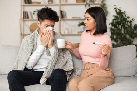 Supportive Asian Wife Treating Sick Husband With Flu Giving Him Medicine Sitting On Couch At Home. Korean Girlfriend Takes Care Of Ill Boyfriend. Seasonal Influenza, Cold, Illness Conceptの写真素材