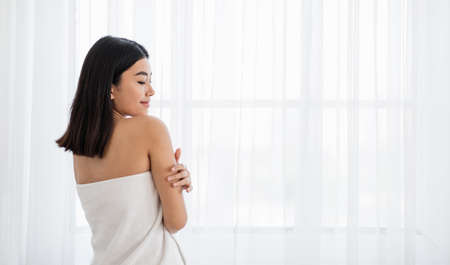 Joyful asian lady in towel touching her skin while standing by window at home. Young woman wrapped in white bath towel enjoying her soft skin after morning shower, panorama with copy spaceの写真素材