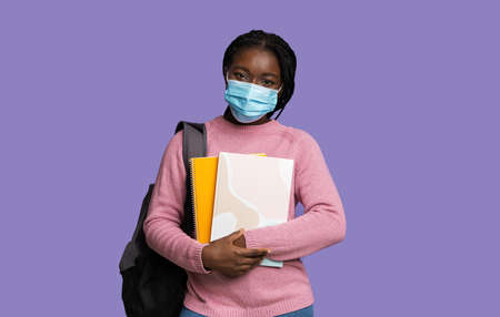 Covid-19 And Education Concept. Black Female University Student Wearing Medical Mask, Holding Workbooks And Backpack, Looking At Camera While Posing Over Purple Studio Background, Copy Spaceの写真素材
