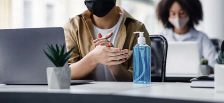 Focus on antiseptic to protect against covid virus. Guy disinfects his hands, sitting at table at workplace with laptop, working in interior of modern office, panorama, copy spaceの写真素材
