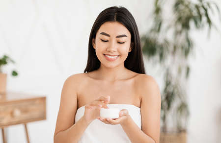 Happy asian woman wrapped in towel using face or body nourishing cream, bedroom interior. Pretty young lady using moisturizer for her face or body after shower in the morning, copy spaceの写真素材