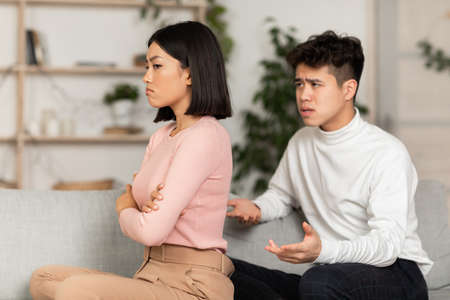 Asian Husband Trying To Talk With Offended Wife After Family Conflict Sitting On Couch At Home....