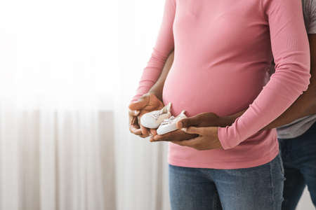 Cropped of unrecognizable expecting black family in casual holding small baby shoes together. African american man and pregnant woman with baby booties standing by window at home, closeup, copy spaceの写真素材