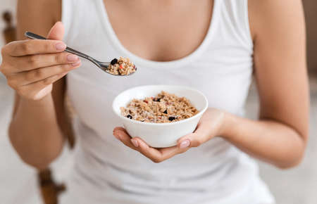 Healthy food and diet concept. Cropped image of young woman holding bowl with homemade granola. Healthy unrecognizable lady eating nutritive oatmeal at home, holding plate and spoon, closeupの写真素材