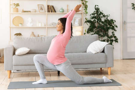 Side view of healthy pregnant black woman exercising alone at home, doing morning yoga, empty space. Active expecting african american lady with big tummy training on fitness mat at living roomの写真素材