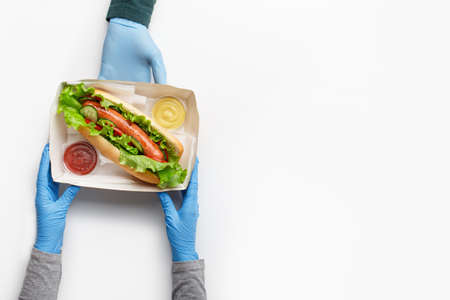 Modern lunch and healthy fast food. Waiter in rubber gloves gives order to customer in cardboard box with big hot dog with vegetables and lettuce with ketchup and sauce in containers isolated on white background, top viewの写真素材