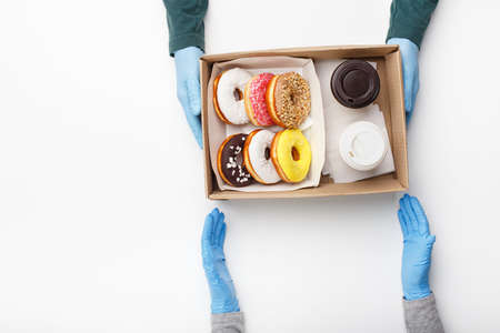 Customer in rubber gloves picks up order during coronavirus outbreak. Waiter gives box with donuts with glaze and two cups of coffee, takeaway, isolated on white background, top view, copy spaceの写真素材