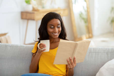 Stay home leisure activities. Lovely young black woman reading book with cup of hot drink in living...