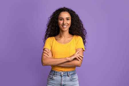 Portrait Of Confident Young Woman With Curly Hair Posing With Folded Arms Over Purple Background In Studio, Beautiful Millennial Lady With Tanned Skin Looking And Smiling At Camera, Copy Spaceの写真素材