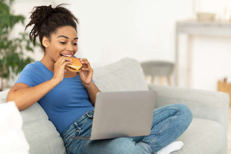 Cheat Meal. Black Woman Enjoying Unhealthy Burger Sitting At Laptop And Watching Movie At Home On...