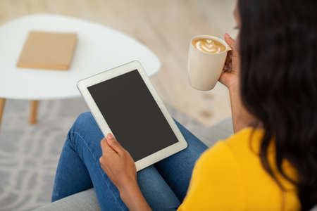 Young black woman with coffee sitting on sofa and using tablet computer with blank screen at home,...