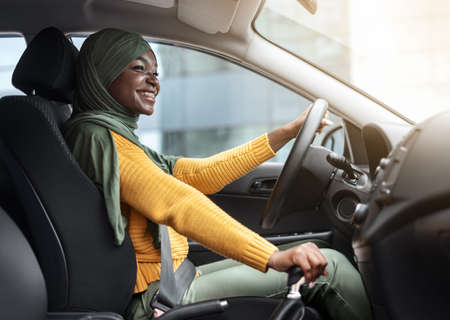 Confident Driver. Smiling African Muslim Woman In Headscarf Driving Modern Car In City, Young Black Islamic Female In Hijab Enjoying Drive New Vehicle, Holding Steering Wheel And Looking On Roadの写真素材