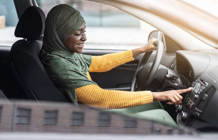 Car Playlist. Joyful Black Muslim Woman In Hijab Driving Car And Listening Music From Auto Audio...