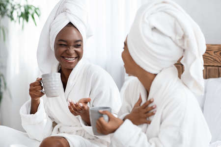 Two joyful african american girlfriends in bathrobes joking and drinking coffee. Cheerful black women with towels on their head having spa day at home, drinking tea and having funny conversationの写真素材